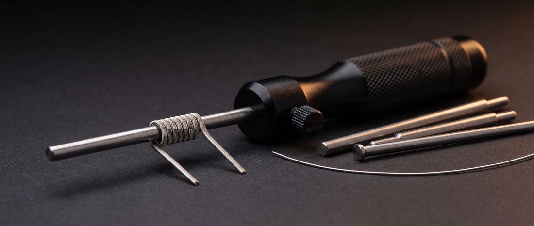 Coil jig with a freshly wrapped vape coil on its mandrel rod, side thumbscrew, three spare wrapping posts and a strand of resistance wire on a dark studio surface