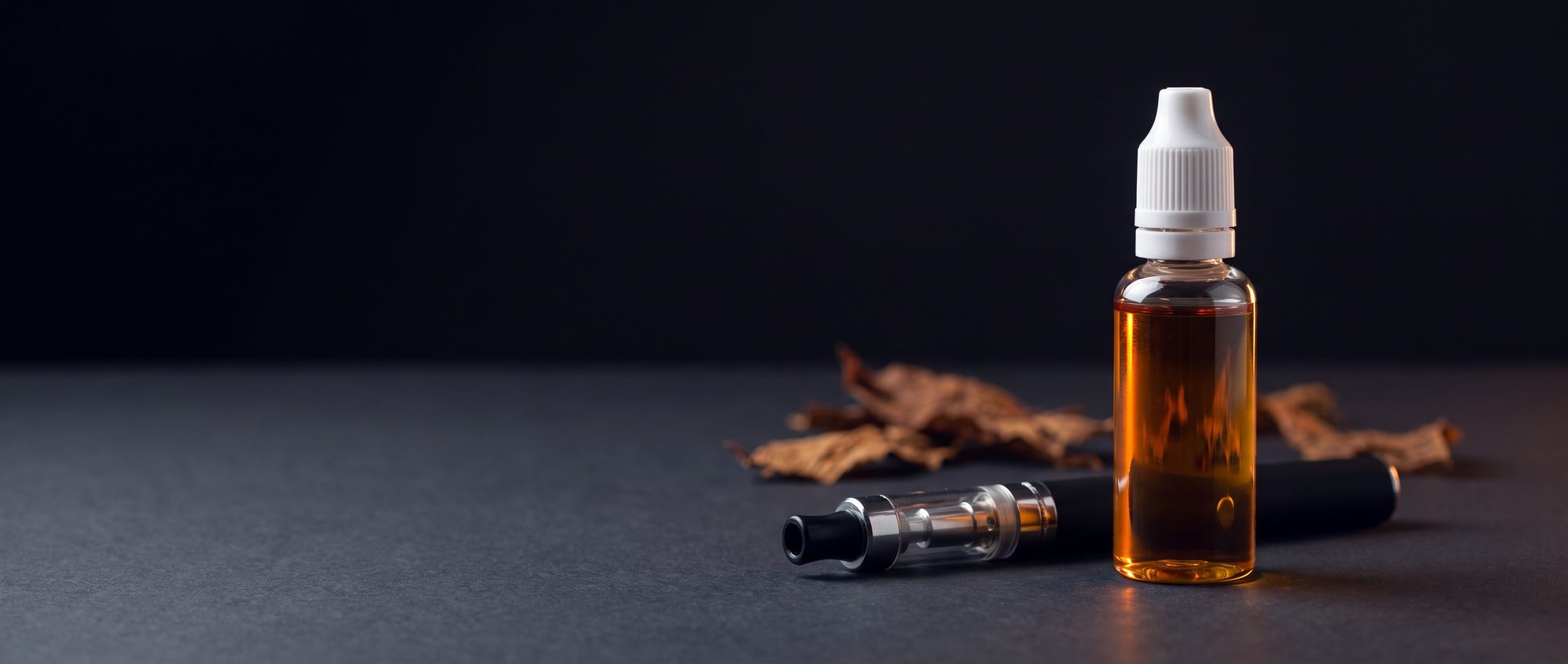 Unlabeled bottle of amber e-liquid beside a classic pen-style vape with a clearomizer and dried tobacco leaves on a dark studio surface