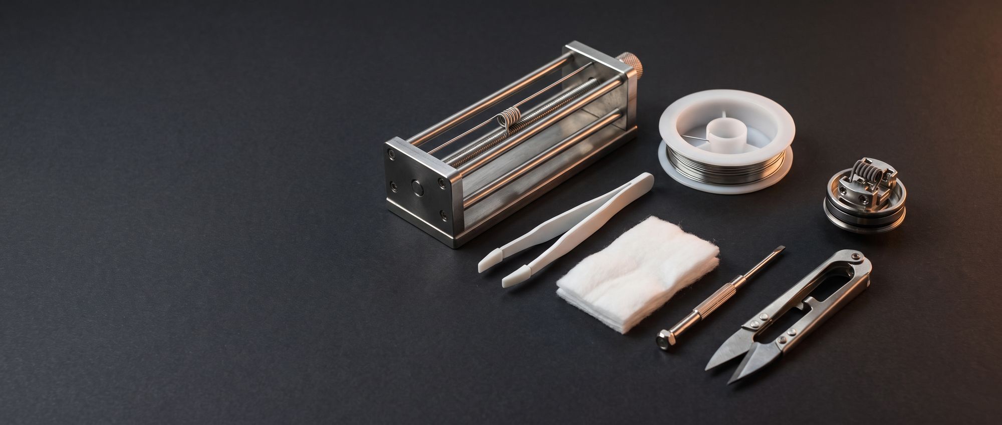 Coil building tools laid out on a dark bench: coil jig, spool of resistance wire, ceramic tweezers, organic cotton, screwdriver, wire snips and a rebuildable atomizer deck with a wrapped coil
