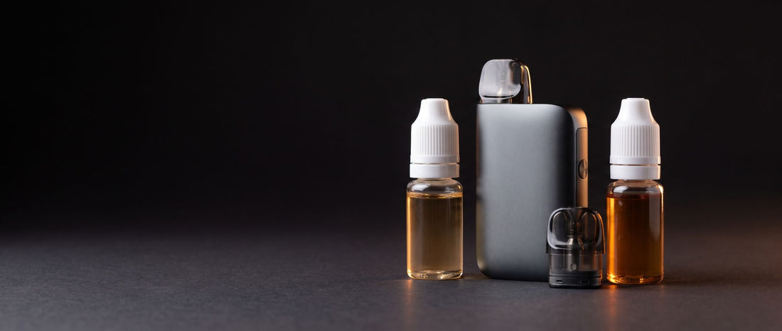 Compact pod vape device with a spare pod between two small bottles of e-liquid, one light and one dark amber, on a dark studio surface
