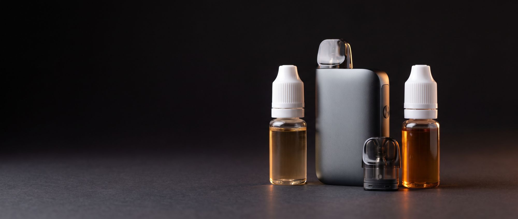 Compact pod vape device with a spare pod between two small bottles of e-liquid, one light and one dark amber, on a dark studio surface