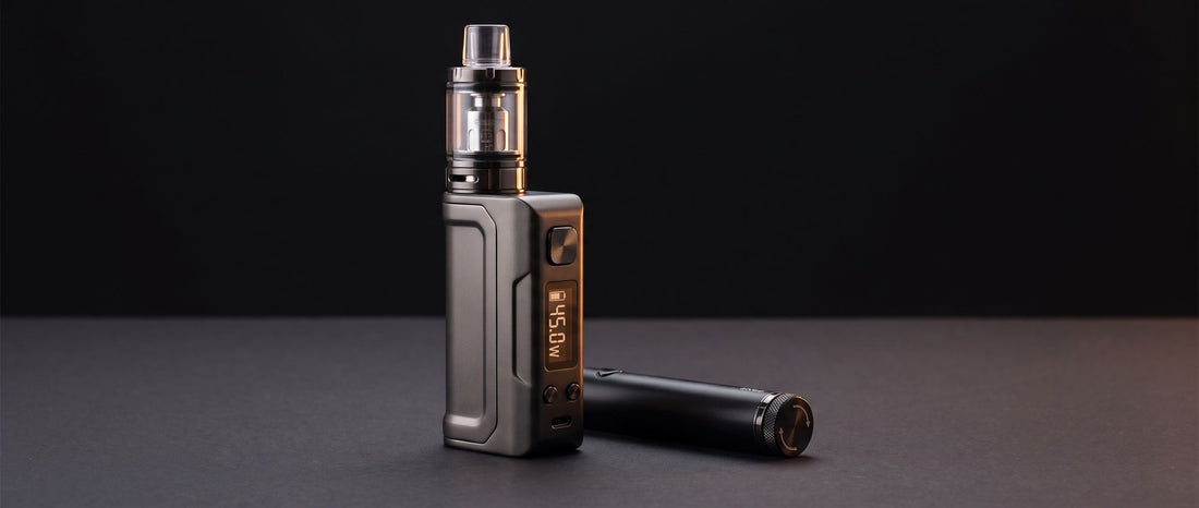 Compact box mod vape with a glass tank and a wattage display next to a slim twist-dial pen battery on a dark studio surface
