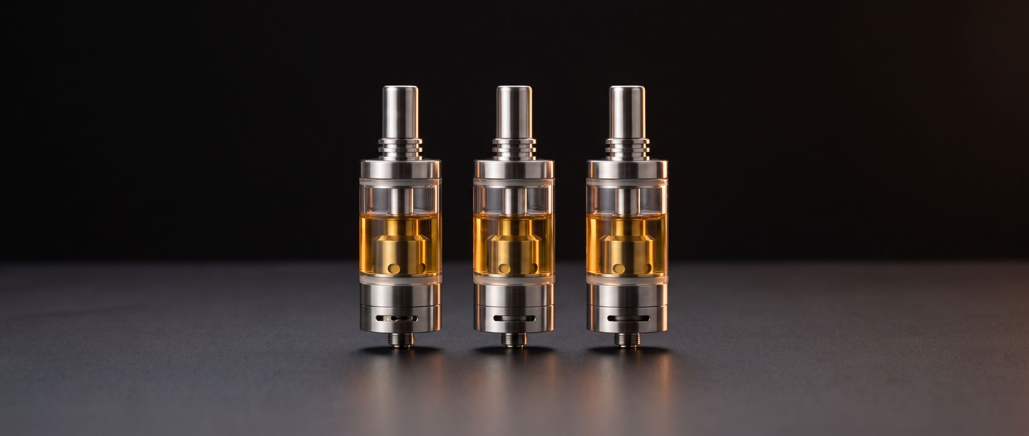 Three pen-style clearomizer tanks with glass bodies and stainless steel bases on a dark studio background