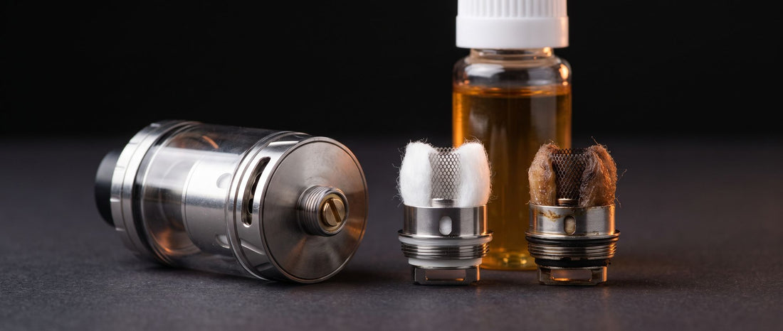 Sub-ohm vape tank lying beside a fresh mesh coil head with clean white cotton and a used coil head with scorched brown cotton, an e-liquid bottle behind them on a dark studio surface