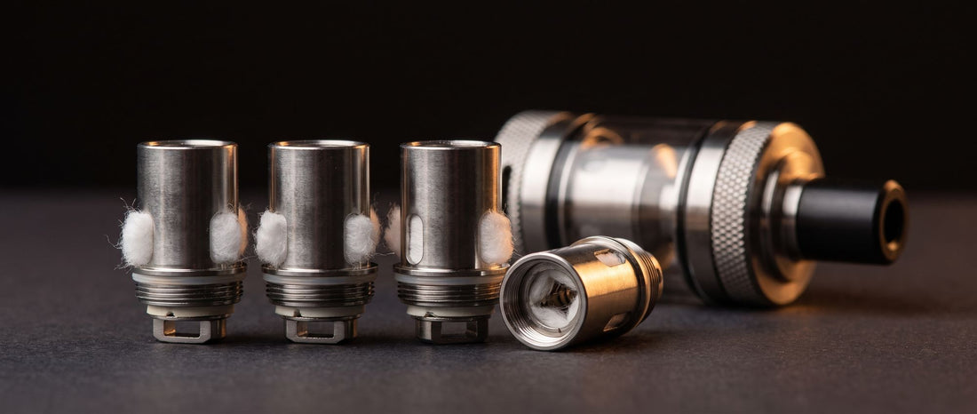 Four stainless steel BVC replacement coil heads with cotton wicking ports in front of a mouth-to-lung vape tank on a dark studio surface