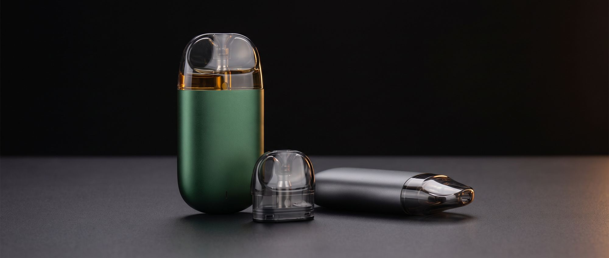 Compact green pod vape standing upright with its clear pod filled, a graphite pod vape lying beside it and a spare pod in front, on a dark studio surface