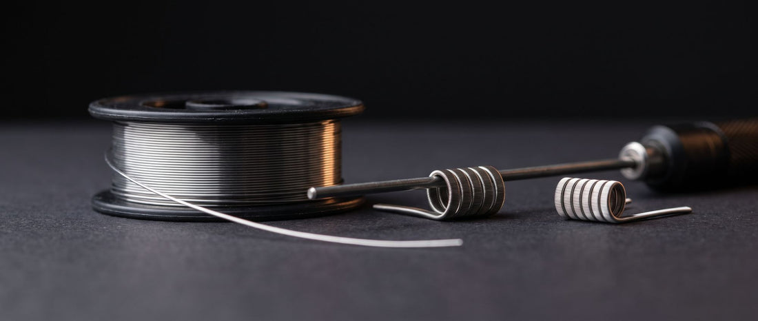 Spool of Kanthal resistance wire beside two wrapped vape coils, one still on a coil jig rod, on a dark studio surface