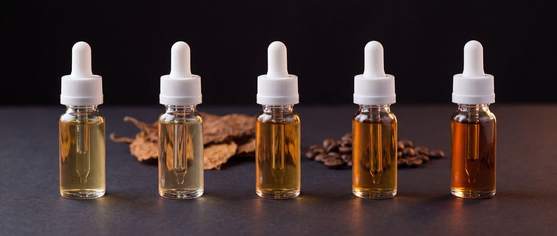 Five unlabeled e-liquid dropper bottles in shades from pale straw to deep amber, with dried tobacco leaves and coffee beans behind them on a dark studio surface