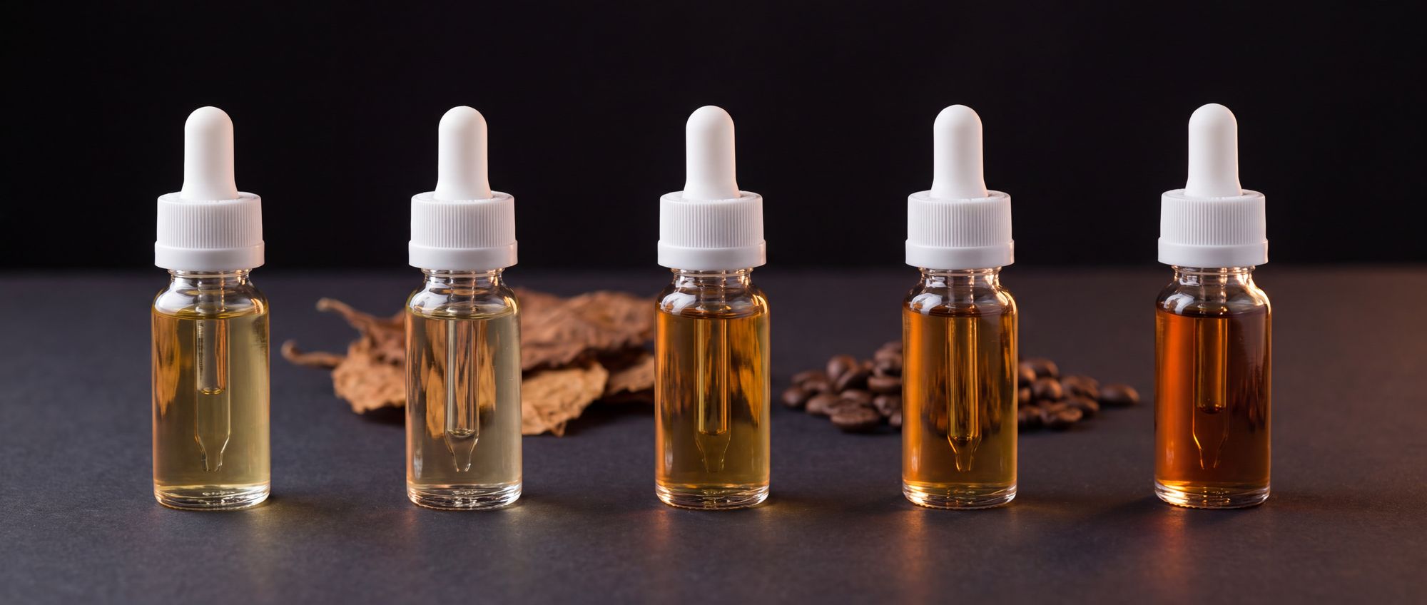 Five unlabeled e-liquid dropper bottles in shades from pale straw to deep amber, with dried tobacco leaves and coffee beans behind them on a dark studio surface